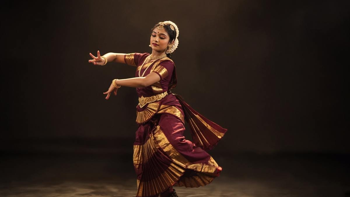 A classical dancer in burgundy and gold, caught mid-movement on a dark stage.